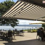 Zdjęcie prezentuje szeroki, zewnętrzny taras restauracyjny położony bezpośrednio nad brzegiem jeziora, w pełni osłonięty przez dwie połączone markizy Corsica. Poszycie markiz posiada klasyczny wzór w szerokie, pionowe pasy w odcieniach beżu i ciemnego brązu, co harmonizuje z naturalnym otoczeniem. Pod zadaszeniem ustawiono liczne stoliki kawowe oraz ciemnobrązowe krzesła z technorattanu. Przy jednym ze stolików siedzą goście, korzystając z cienia w słoneczny dzień. Taras jest wyłożony szarą kostką brukową i oddzielony od jeziora niskim, metalowym ogrodzeniem panelowym. W tle rozpościera się widok na spokojną taflę wody, na której unoszą się małe łodzie, oraz na zarysy gór na horyzoncie pod jasnym niebem. Po lewej stronie tarasu rośnie rozłożyste, zielone drzewo, przy którym stoi złożony parasol ogrodowy w kolorze kremowym. W centralnej części placu widoczny jest również drugi, rozstawiony biały parasol reklamowy. Całość sceny ukazuje funkcjonalne zastosowanie dużych osłon przeciwsłonecznych w branży gastronomicznej, tworzących komfortowe miejsce wypoczynku z widokiem na naturę.