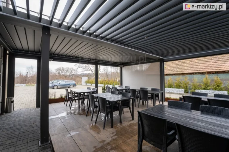 Czarne prostokątne stoliki z krzesłami, ustawione na drewnianym podeście przed wejściem restauracji zadaszone pergolą SB400 Selt, który zapewnia ochronę przed warunkami atmosferycznymi, jednocześnie tworząc elegancką przestrzeń na świeżym powietrzu. Ilość światła kontrolowana jest przez uchylenie lamele oraz boczne osłony refleksol Ziiip 120 Selt.