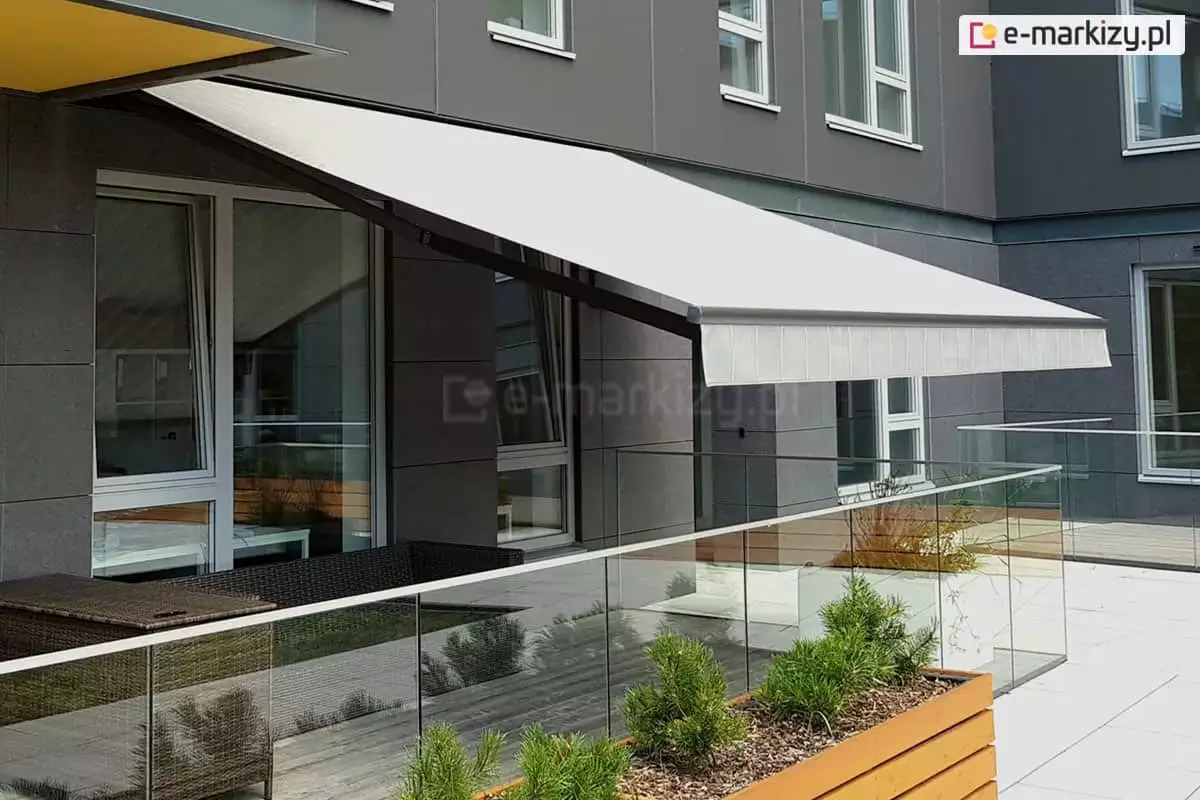 Markiza Jamaica Selt z prostą falbaną zamocowana na fasadzie budynku z elewacją w odcieniach szarości. Markiza o dużym kącie nachylenia zacienia duże okna tarasowe oraz przestrzeń tarasu wyznaczoną przez szklane barierki i zapewnia cień ustawionym na tarasie meblom z technorattanu. Przed barierką tarasową ustawiona drewniana donica z malutkimi iglakami.