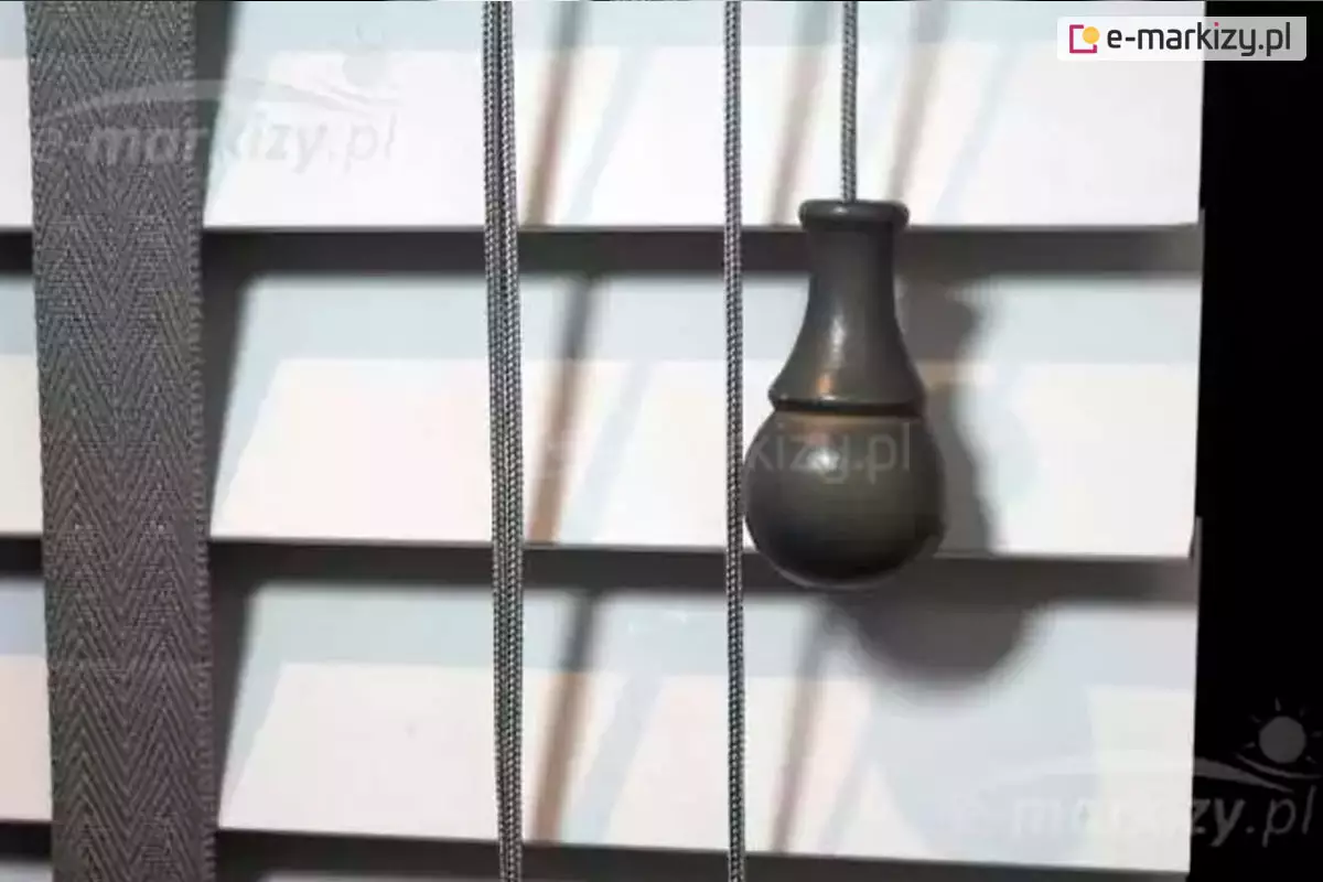 Żaluzja drewniana z lamelami 50 mm w kolorze białym z pasmanterią i drabinką taśmową w kolorze grafitowym. Widoczny także ozdobny obciążnik sznurka do sterowania lamelami.