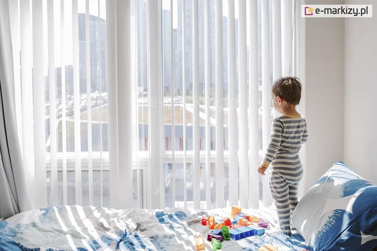 Jasne wnętrze sypialni lub pokoju dziecięcego, w którym duże okno zostało osłonięte przez białe żaluzje pionowe. Lamele wykonane z półtransparentnego materiału są ustawione prostopadle do szyby, dzięki czemu do pomieszczenia wpada bardzo dużo miękkiego, rozproszonego światła. Przez żaluzje pionowe widać zarys nowoczesnej zabudowy miejskiej w oddali. Na pierwszym planie znajduje się łóżko przykryte pościelą w biało-niebieskie wzory, na której leżą kolorowe klocki do zabawy. Małe dziecko w pasiastej piżamie stoi tyłem do obiektywu na łóżku i wygląda przez okno, trzymając dłoń w pobliżu lameli. Światło przenikające przez żaluzje pionowe tworzy na pościeli delikatne, pasiaste cienie, podkreślając przytulny i bezpieczny charakter domowej przestrzeni. Całość kompozycji jest utrzymana w bardzo jasnej, wysokiej tonacji, co uwydatnia czystość i lekkość zastosowanych osłon okiennych.