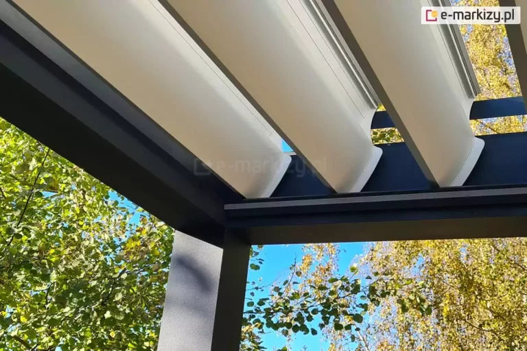 Zdjęcie przedstawia zbliżenie na detale konstrukcyjne dachu pergoli bioklimatycznej w miejscu styku ruchomych lameli z ramą nośną. Szerokie, aluminiowe lamele w kolorze białym są ustawione pod skosem w pozycji półotwartej, co pozwala na regulację przepływu powietrza i światła. Widoczne są precyzyjnie wykonane zakończenia lameli oraz ich osadzenie na czarnych prowadnicach zintegrowanych z masywną, antracytową belką konstrukcyjną. Belka ta charakteryzuje się matowym wykończeniem i prostym, geometrycznym profilem. W tle kadru widać gęste korony drzew liściastych o zielono-żółtym zabarwieniu oraz czyste, błękitne niebo, co wskazuje na słoneczny dzień. Perspektywa od dołu podkreśla techniczną estetykę systemu oraz stabilność aluminiowego szkieletu.