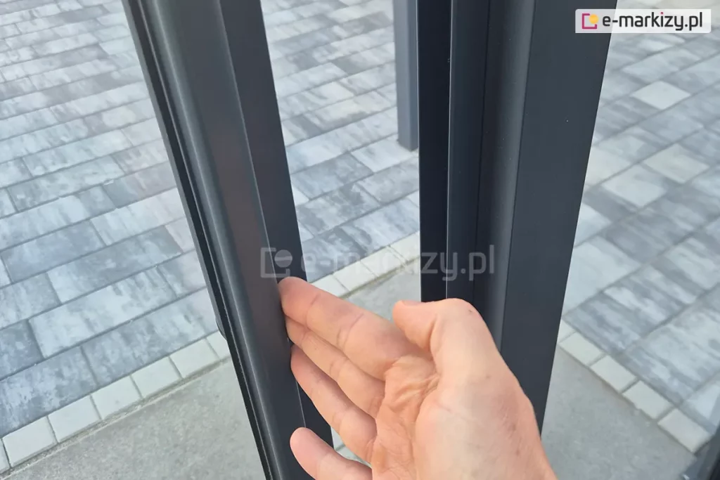Zbliżenie na detal systemu lekkich szklanych drzwi przesuwnych w ramkach aluminiowych. Zdjęcie prezentuje mechanizm otwierania, gdzie dłoń wskazuje na pionowy profil skrzydła przesuwnego wykonany z aluminium malowanego proszkowo na kolor antracytowy. Widoczna jest precyzyjna szczelina między skrzydłami oraz system uszczelnień. Konstrukcja ramy charakteryzuje się nowoczesnym, minimalistycznym designem. W tle, pod dolną prowadnicą, widoczna jest nawierzchnia tarasu wykonana z jasnoszarej kostki brukowej o prostokątnym kształcie, ułożonej w regularny wzór. Ostrość zdjęcia skoncentrowana jest na krawędzi profilu, podkreślając jakość wykończenia elementów zabudowy.