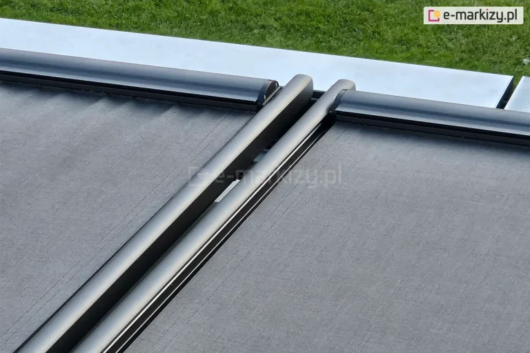 Zdjęcie przedstawia techniczne zbliżenie na detale konstrukcyjne systemu veranda w wesji modułowej. Na pierwszym planie widoczne są dwie równoległe prowadnice boczne oraz profile czołowe, lakierowane proszkowo na kolor ciemnego antracytu o matowym wykończeniu. Między profilami rozpięta jest wysokiej jakości tkanina techniczna w kolorze grafitowym o wyraźnej strukturze splotu. Konstrukcja charakteryzuje się nowoczesnym, minimalistycznym designem z zaokrąglonymi krawędziami profili aluminiowych. W górnej części kadru widoczny jest fragment jasnej, metalowej obróbki blacharskiej oraz pas zielonego trawnika w tle, co wskazuje na montaż systemu na zadaszeniu ogrodu zimowego lub pergoli. Zdjęcie kładzie nacisk na precyzję spasowania elementów mechanicznych i estetykę wykończenia systemu osłonowego.