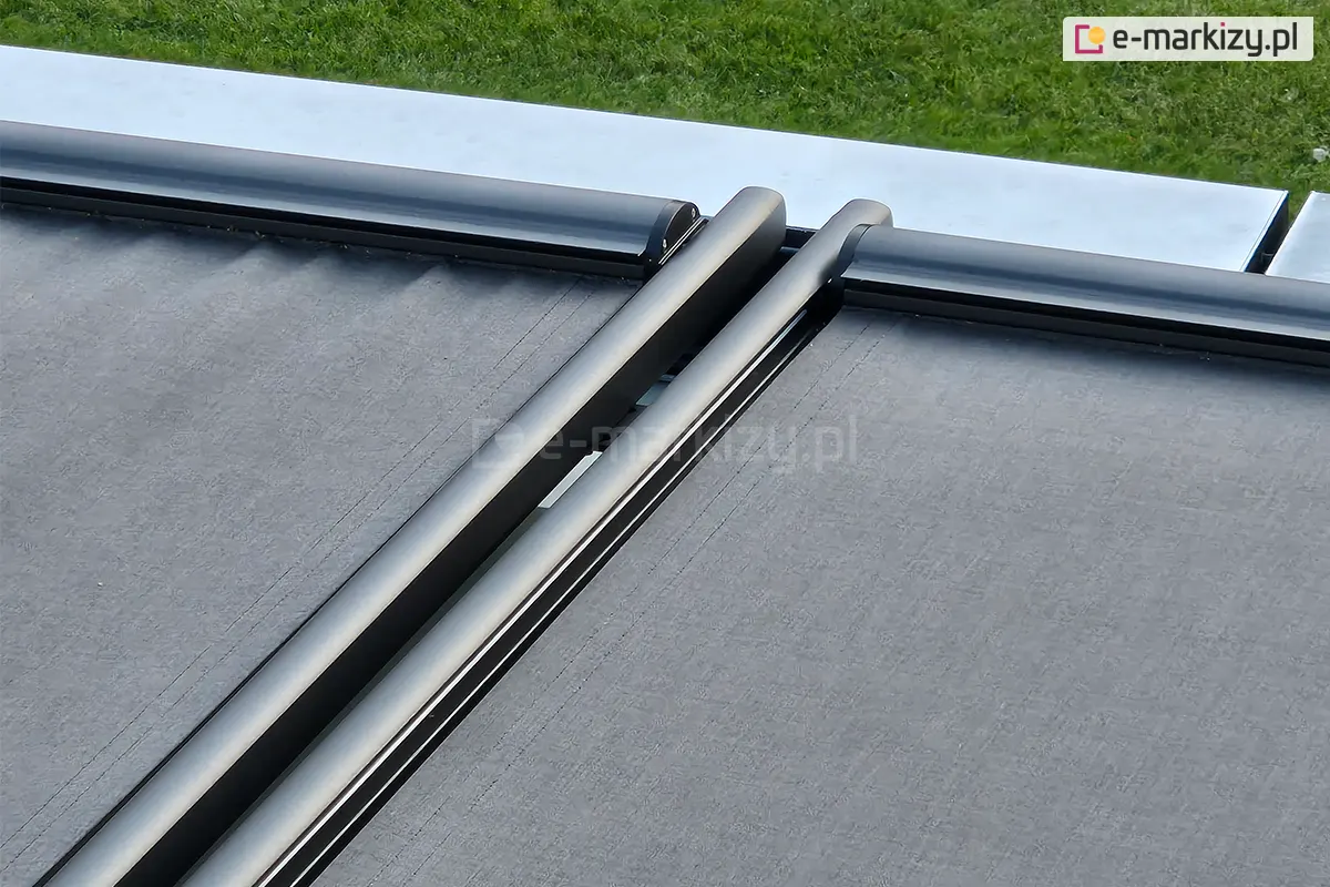 Zdjęcie przedstawia techniczne zbliżenie na detale konstrukcyjne systemu veranda w wesji modułowej. Na pierwszym planie widoczne są dwie równoległe prowadnice boczne oraz profile czołowe, lakierowane proszkowo na kolor ciemnego antracytu o matowym wykończeniu. Między profilami rozpięta jest wysokiej jakości tkanina techniczna w kolorze grafitowym o wyraźnej strukturze splotu. Konstrukcja charakteryzuje się nowoczesnym, minimalistycznym designem z zaokrąglonymi krawędziami profili aluminiowych. W górnej części kadru widoczny jest fragment jasnej, metalowej obróbki blacharskiej oraz pas zielonego trawnika w tle, co wskazuje na montaż systemu na zadaszeniu ogrodu zimowego lub pergoli. Zdjęcie kładzie nacisk na precyzję spasowania elementów mechanicznych i estetykę wykończenia systemu osłonowego.