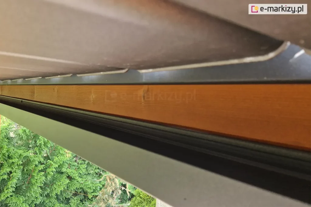 Zbliżenie systemu rynnowego zamocowanego do konstrukcji drewnianej wzdłuż wysięgu dachu lamelowego SB400 MD Selt. Pod dachem widoczne zielone iglaki.