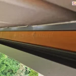 Zbliżenie systemu rynnowego zamocowanego do konstrukcji drewnianej wzdłuż wysięgu dachu lamelowego SB400 MD Selt. Pod dachem widoczne zielone iglaki.