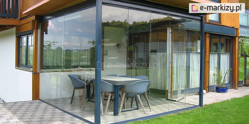 Nowoczesna zabudowa narożna wykonana z bezramowych, przesuwnych szyb szklanych Miros, zamontowana przy budynku jednorodzinnym o elewacji łączącej białe i drewnopodobne wykończenia. Przeszklona przestrzeń tarasu oparta jest na aluminiowej konstrukcji w kolorze antracytowym i obejmuje dwie ściany boczne. Na podłożu ułożona została posadzka z płytek ceramicznych w szachownicowy wzór w odcieniach szarości. Wewnątrz zabudowy znajduje się nowoczesny stół jadalniany z tworzywa sztucznego oraz sześć ergonomicznych krzeseł. Zabudowa zapewnia pełną widoczność na zewnątrz – w tle widoczne są tereny zielone, drzewa i fragment zabudowy sąsiadującej. Obok znajduje się taras i trawnik z przystrzyżoną trawą, a całość jest częścią ogrodu przy nowoczesnym domu.