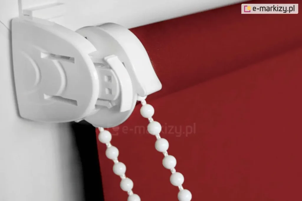 Zbliżenie na uchwyt i mechanizm rolety mini wykonane w kolorze białym z dopasowanym kolorystycznie łańcuszkiem do sterowania roletką, która wyposażona została w tkaninę o pięknym czerwonym kolorze.
