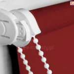 Zbliżenie na uchwyt i mechanizm rolety mini wykonane w kolorze białym z dopasowanym kolorystycznie łańcuszkiem do sterowania roletką, która wyposażona została w tkaninę o pięknym czerwonym kolorze.