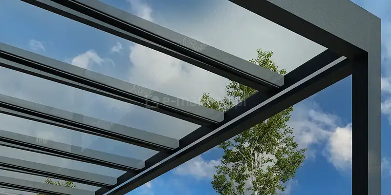 Detal techniczny systemu zadaszenia szklanego Spekta Glass, ukazany z perspektywy od dołu na tle błękitnego nieba z lekkimi chmurami. Konstrukcja nośna została wykonana z grubych profili aluminiowych w kolorze antracytowym, które tworzą rytmiczny układ krokwi podtrzymujących szklane panele. Panele dachowe są wykonane z bezpiecznego, w pełni przeziernego szkła, co zapewnia nieograniczony widok na niebo i maksymalny dostęp światła słoneczno-dziennego. Profile aluminiowe charakteryzują się prostymi liniami i matowym wykończeniem, co podkreśla nowoczesny, industrialny charakter systemu. W tle, za krawędzią zadaszenia, widoczna jest korona zielonego drzewa, co ilustruje, jak szklana konstrukcja harmonijnie przenika się z otaczającą naturą, nie zasłaniając widoku.