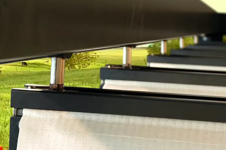 Prowadnica dachu pergoli Dragon z widocznymi zawiesiami ze stali, w których mocowane są antracytowe belki przeciwwietrzne. Pomiędzy belkami widoczna biała dziurkowana tkanina zadaszenia, a w tle zielone łąki.