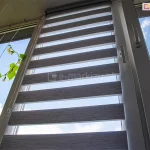 Szare rolety okienne dzień i noc zamontowane na oknie wyposażonym także w moskitierę z kwiatami rosnącymi obok.