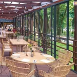 Szklane ściany przesuwne ramowe Sotro zastosowane jako osłona zadaszonego tarasu w restauracji. Przesuwne panele szklane zapewniają ochronę przed wiatrem i deszczem, a jednocześnie pozwalają wpuścić na taras świeże powietrze. Pod zadaszeniem na drewnianym tarasie ustawiono kilka różnych stolików z krzesłami dla klientów. Pod zadaszeniem widoczne także sznury z lampkami. Za przeszkleniem widoczny zielony trawnik oraz drzewa ocieniające ścieżkę.