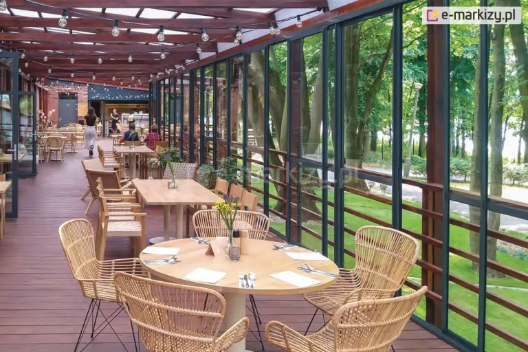 Szklane ściany przesuwne ramowe Sotro zastosowane jako osłona zadaszonego tarasu w restauracji. Przesuwne panele szklane zapewniają ochronę przed wiatrem i deszczem, a jednocześnie pozwalają wpuścić na taras świeże powietrze. Pod zadaszeniem na drewnianym tarasie ustawiono kilka różnych stolików z krzesłami dla klientów. Pod zadaszeniem widoczne także sznury z lampkami. Za przeszkleniem widoczny zielony trawnik oraz drzewa ocieniające ścieżkę.