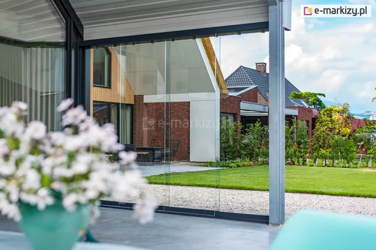 Widok przez panoramiczne, bezramowe szklane ściany przesuwne systemu Auris, zamontowane na nowoczesnym tarasie. Zdjęcie prezentuje wysoką transparentność tafli szkła hartowanego, które odbijają architekturę sąsiedniego budynku o nowoczesnej bryle z dachem krytym strzechą i elewacją z czerwonej cegły. Konstrukcja wsporcza w kolorze szarym/antracytowym tworzy ramę dla widoku na zadbany, zielony trawnik oraz niską roślinność ogrodową. Na pierwszym planie, wewnątrz zabudowy, widoczne są rozmyte białe kwiaty w donicy, co podkreśla głębię obrazu. System przesuwny Auris pozwala na płynne połączenie wnętrza domu z przestrzenią ogrodu, zapewniając ochronę przed wiatrem przy zachowaniu pełnej jasności pomieszczenia.