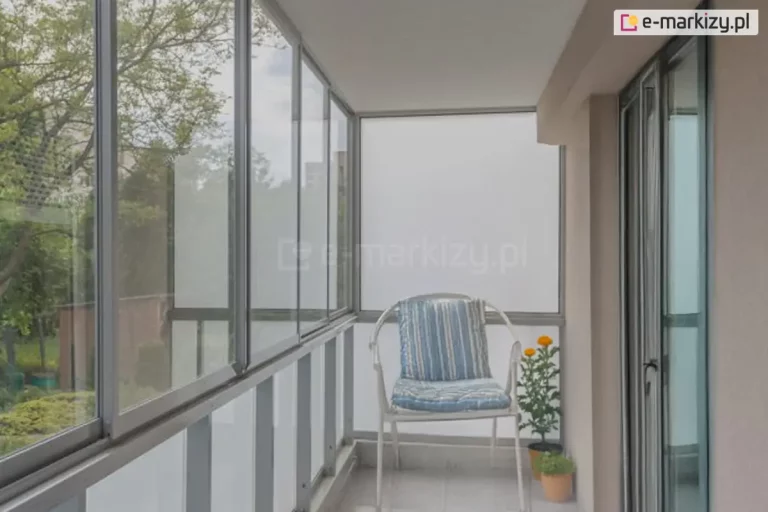 Przesuwna szklana zabudowa balkonu wykonana w lekkim systemie aluminiowym z profilami w kolorze srebrnym (naturalne aluminium). Konstrukcja składa się z szeregu pionowych, szklanych paneli osadzonych w ramkach, które poruszają się po wielotorowej prowadnicy zamontowanej na balustradzie balkonowej. Boczna sekcja zabudowy została wypełniona nieprzeziernym szkłem matowym (mlecznym), zapewniającym prywatność użytkownikom. Na balkonie widoczne jest białe krzesło ogrodowe z niebiesko-białą poduszką oraz małe doniczki z roślinami. System jest zamontowany do sufitu i bocznej ściany budynku, tworząc szczelną barierę chroniącą przed wiatrem i deszczem, przy zachowaniu widoku na zieleń za oknem.