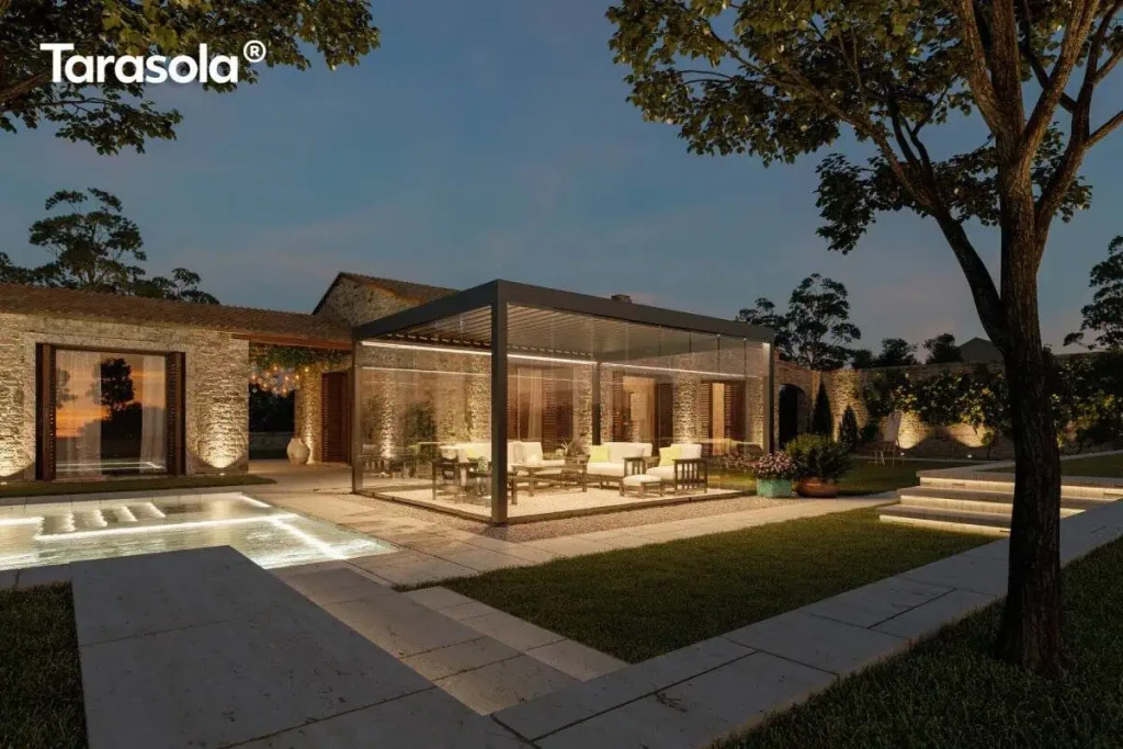 Nowoczesna pergola lamelowa essential Tarasola w kolorze ciemnoantracytowym, przedstawiona w nastrojowej scenerii wieczornej z włączonym oświetleniem taśmami LED zamontowanymi wzdłuż ramy dachu. Konstrukcja posiada pełną szklaną zabudowę z przesuwnymi panelami, które tworzą zamknięty, transparentny pawilon ogrodowy oświetlający wnętrze strefy wypoczynkowej. W środku, na podłożu z dekoracyjnego żwiru, znajduje się zestaw luksusowych mebli tarasowych z jasnymi poduszkami. Pergola jest usytuowana na tarasie z jasnych płyt kamiennych obok basenu, którego wnętrze jest podświetlone pod wodą. W tle widoczny jest parterowy budynek z elewacją z naturalnego kamienia oraz kamienny mur, oba oświetlone punktowymi reflektorami podkreślającymi ich strukturę. Całość aranżacji dopełnia zadbany trawnik, kamienne schody oraz drzewa, tworząc elegancką przestrzeń rekreacyjną pod ciemniejącym, wieczornym niebem.