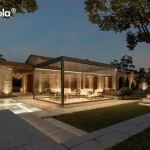 Nowoczesna pergola lamelowa essential Tarasola w kolorze ciemnoantracytowym, przedstawiona w nastrojowej scenerii wieczornej z włączonym oświetleniem taśmami LED zamontowanymi wzdłuż ramy dachu. Konstrukcja posiada pełną szklaną zabudowę z przesuwnymi panelami, które tworzą zamknięty, transparentny pawilon ogrodowy oświetlający wnętrze strefy wypoczynkowej. W środku, na podłożu z dekoracyjnego żwiru, znajduje się zestaw luksusowych mebli tarasowych z jasnymi poduszkami. Pergola jest usytuowana na tarasie z jasnych płyt kamiennych obok basenu, którego wnętrze jest podświetlone pod wodą. W tle widoczny jest parterowy budynek z elewacją z naturalnego kamienia oraz kamienny mur, oba oświetlone punktowymi reflektorami podkreślającymi ich strukturę. Całość aranżacji dopełnia zadbany trawnik, kamienne schody oraz drzewa, tworząc elegancką przestrzeń rekreacyjną pod ciemniejącym, wieczornym niebem.