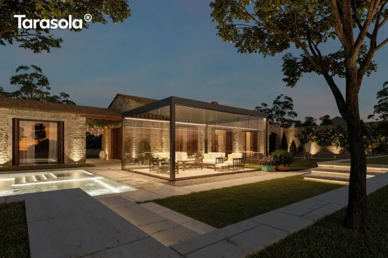 Nowoczesna pergola lamelowa essential Tarasola w kolorze ciemnoantracytowym, przedstawiona w nastrojowej scenerii wieczornej z włączonym oświetleniem taśmami LED zamontowanymi wzdłuż ramy dachu. Konstrukcja posiada pełną szklaną zabudowę z przesuwnymi panelami, które tworzą zamknięty, transparentny pawilon ogrodowy oświetlający wnętrze strefy wypoczynkowej. W środku, na podłożu z dekoracyjnego żwiru, znajduje się zestaw luksusowych mebli tarasowych z jasnymi poduszkami. Pergola jest usytuowana na tarasie z jasnych płyt kamiennych obok basenu, którego wnętrze jest podświetlone pod wodą. W tle widoczny jest parterowy budynek z elewacją z naturalnego kamienia oraz kamienny mur, oba oświetlone punktowymi reflektorami podkreślającymi ich strukturę. Całość aranżacji dopełnia zadbany trawnik, kamienne schody oraz drzewa, tworząc elegancką przestrzeń rekreacyjną pod ciemniejącym, wieczornym niebem.