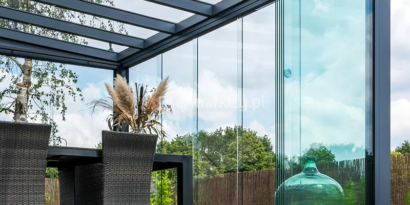Wnętrze nowoczesnego ogrodu letniego opartego na systemie zadaszenia Terano. Głównym elementem kadru jest narożna sekcja szklanej zabudowy, składająca się z bezramowych paneli przesuwnych wykonanych z hartowanego szkła. Panele są zamontowane w subtelnych szynach, a na jednej z tafli widoczny jest minimalistyczny, okrągły uchwyt do przesuwania. Konstrukcja wsporcza, w tym krokwie dachowe oraz słupy narożne, wykonana jest z aluminium lakierowanego proszkowo na kolor ciemnego antracytu. Pod zadaszeniem widoczny jest fragment stołu jadalnianego z krzesłami o splotach technorattanowych w kolorze szarym. Na stole ustawiono dekoracyjną wazę z suszonymi trawami pampasowymi, a obok, na podłodze, stoi duży szklany gąsior w kolorze morskiej zieleni. Przez transparentne ściany i dach widać korony drzew, brzozę oraz zachmurzone niebo, co podkreśla efekt zatarcia granicy między wnętrzem a ogrodem. W oddali dostrzec można również naturalne ogrodzenie wykonane z mat wiklinowych lub bambusowych.