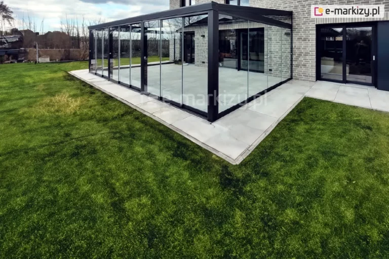 Nowoczesna, narożna zabudowa tarasu przy budynku mieszkalnym, wykonana z czarnych profili aluminiowych i przezroczystych, bezramowych paneli szklanych Miros. Konstrukcja osadzona na jasnoszarej, betonowej posadzce, ściśle przylega do elewacji z szarej cegły. Ściany przesuwne umożliwiają otwieranie przestrzeni na ogród. Na pierwszym planie rozciąga się równy, intensywnie zielony trawnik. W tle widoczna zabudowa jednorodzinna, nagie drzewa i zachmurzone, niebiesko-szare niebo.