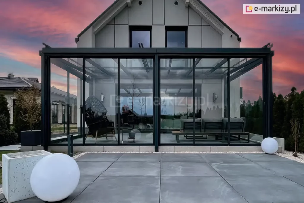 Kompleksowa szklana zabudowa tarasu w systemie ramowym, zintegrowana z nowoczesną bryłą domu jednorodzinnego. Zdjęcie przedstawia szerokie zadaszenie tarasu oparte na ciemnych profilach aluminiowych, wypełnione hartowanym szkłem. Front zabudowy składa się z wieloskrzydłowego systemu drzwi przesuwnych w cienkich ramkach, podzielonego na dwie sekcje. Wewnątrz przeszklonego ogrodu zimowego widoczne są meble wypoczynkowe, w tym stylowy fotel oraz sofa. Konstrukcja posiada szklany dach wsparty na krokwiach aluminiowych. Przed zabudową znajduje się taras z dużych, szarych płyt betonowych, ozdobiony nowoczesnymi lampami ogrodowymi w kształcie białych kul. Tło stanowi elewacja budynku z nowoczesnych paneli oraz niebo w kolorach zachodu słońca.