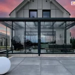 Kompleksowa szklana zabudowa tarasu w systemie ramowym, zintegrowana z nowoczesną bryłą domu jednorodzinnego. Zdjęcie przedstawia szerokie zadaszenie tarasu oparte na ciemnych profilach aluminiowych, wypełnione hartowanym szkłem. Front zabudowy składa się z wieloskrzydłowego systemu drzwi przesuwnych w cienkich ramkach, podzielonego na dwie sekcje. Wewnątrz przeszklonego ogrodu zimowego widoczne są meble wypoczynkowe, w tym stylowy fotel oraz sofa. Konstrukcja posiada szklany dach wsparty na krokwiach aluminiowych. Przed zabudową znajduje się taras z dużych, szarych płyt betonowych, ozdobiony nowoczesnymi lampami ogrodowymi w kształcie białych kul. Tło stanowi elewacja budynku z nowoczesnych paneli oraz niebo w kolorach zachodu słońca.