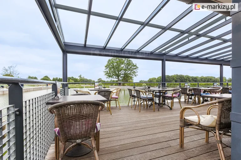 Systemowa szklana zabudowa tarasu w obiekcie komercyjnym, oparta na aluminiowej konstrukcji zadaszenia modelu Terano w kolorze antracytowym. Przeszklony dach o konstrukcji jednospadowej składa się z licznych szklanych paneli osadzonych na aluminiowych krokwiach, wspartych na solidnych słupach nośnych. Przestrzeń tarasu jest otwarta na boki, zabezpieczona metalową balustradą o strukturze kratownicy z poręczą. Podłoże tarasu wykonane jest z ryflowanych desek kompozytowych w naturalnym odcieniu brązu. Wyposażenie obejmuje liczne stoliki restauracyjne z ciemnymi blatami oraz plecione fotele technorattanowe z kolorowymi poduszkami dekoracyjnymi. Taras usytuowany jest nad brzegiem zbiornika wodnego, a w tle widoczny jest rozległy teren zielony z drzewami pod zachmurzonym niebem.