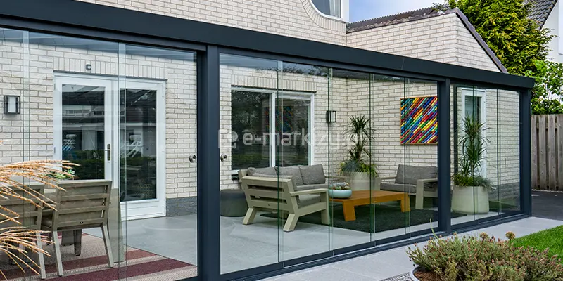 Szklana zabudowa tarasu oparta na systemie Terano, która tworzy zamknięty ogród letni. Konstrukcja wykonana jest z profili aluminiowych w kolorze antracytowym, zamontowanych do elewacji budynku z jasnej, niemal białej cegły. Front oraz boki zadaszenia zostały wypełnione bezramowymi panelami szklanymi w systemie przesuwnym, wyposażonymi w okrągłe uchwyty ułatwiające przesuwanie tafli. Wewnątrz przeszklonej przestrzeni znajduje się strefa wypoczynkowa z nowoczesnymi meblami o jasnoszarych stelażach i grubych poduszkach, ustawionymi wokół niskiego, drewnianego stolika kawowego. Wnętrze zdobią wysokie rośliny w jasnych donicach oraz kolorowy obraz na ścianie. Po lewej stronie, za szklaną barierą, widoczny jest fragment strefy jadalnianej z drewnianym krzesłem ustawionym na pasiastym dywanie zewnętrznym. Podłoga tarasu wyłożona jest gładkimi, szarymi płytami, a przed konstrukcją widoczny jest pas jasnych płyt gresowych oraz fragment trawnika i roślinności ogrodowej.