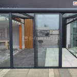 Szklany system drzwi przesuwnych ramowych zamontowany w nowoczesnej, antracytowej konstrukcji zadaszenia tarasowego. Zdjęcie przedstawia dwa rozwiązania osłony bocznej: lewy panel stały w ramie Fest oraz trzy środkowe panele przesuwne Sotro, które zostały zsunięte pośrodku przejścia, ukazując bezprogową szynę dolną. Konstrukcja wykonana z profili aluminiowych malowanych proszkowo na kolor ciemnoszary (antracyt). W tle widoczna podłoga tarasowa wykonana z dużych gresowych płyt w dwóch odcieniach szarości oraz fragment drewnianej pergoli i ściany z pionowymi lamelami dekoracyjnymi.