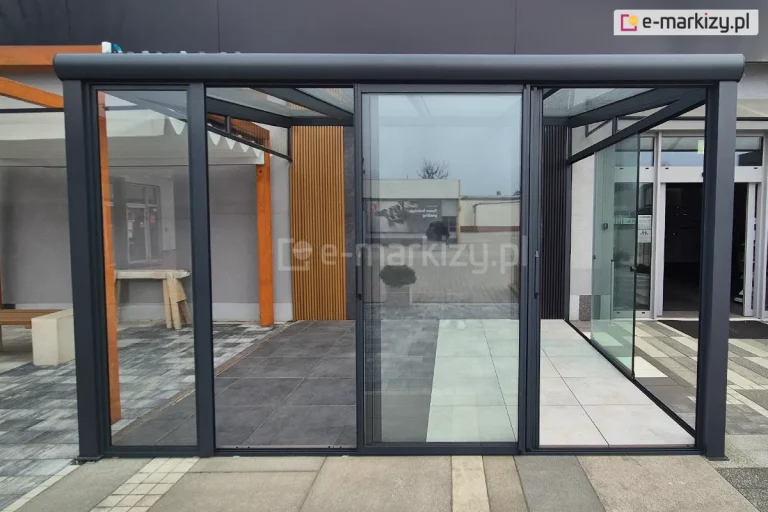 Szklany system drzwi przesuwnych ramowych zamontowany w nowoczesnej, antracytowej konstrukcji zadaszenia tarasowego. Zdjęcie przedstawia dwa rozwiązania osłony bocznej: lewy panel stały w ramie Fest oraz trzy środkowe panele przesuwne Sotro, które zostały zsunięte pośrodku przejścia, ukazując bezprogową szynę dolną. Konstrukcja wykonana z profili aluminiowych malowanych proszkowo na kolor ciemnoszary (antracyt). W tle widoczna podłoga tarasowa wykonana z dużych gresowych płyt w dwóch odcieniach szarości oraz fragment drewnianej pergoli i ściany z pionowymi lamelami dekoracyjnymi.