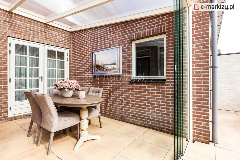 Widok wnętrza przytulnego, przeszklonego tarasu Bosco (nazwa handlowa Lunaro), zaaranżowanego na tle ściany z czerwonej cegły. Konstrukcja posiada jednospadowy dach z półprzezroczystymi panelami poliwęglanu komorowego, osadzonymi w kremowych profilach. Boczną osłonę stanowią bezramowe, szklane panele przesuwne, które są częściowo zsunięte po prawej stronie. W centralnej części tarasu, na podłożu z jasnych płytek ceramicznych, ustawiono okrągły, drewniany stół na ozdobnej, jasnej nodze, przy którym stoją cztery tapicerowane krzesła w kolorze beżowym. Na stole znajduje się duża donica z różowymi kwiatami oraz dwa dekoracyjne lampiony. Ścianę budynku zdobi obraz przedstawiający nadmorski krajobraz, zawieszony obok białego okna i białych, przeszklonych drzwi tarasowych ze szprosami. Po prawej stronie, za szklaną ścianą, widoczny jest pionowy fragment rynny oraz jasne przejście na dalszą część posesji.