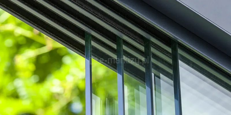 Górna prowadnica szklanych ścian przesuwnych Auris w kolorze antracytowym zamocowana do konstrukcji o tym samym kolorze. W prowadnicy widoczne zamocowane cztery panele szklane o grubości 10mm. W torach przy szybie widoczne uszczelki szczotkowe. W tle rozmyte zielone liście drzewa.