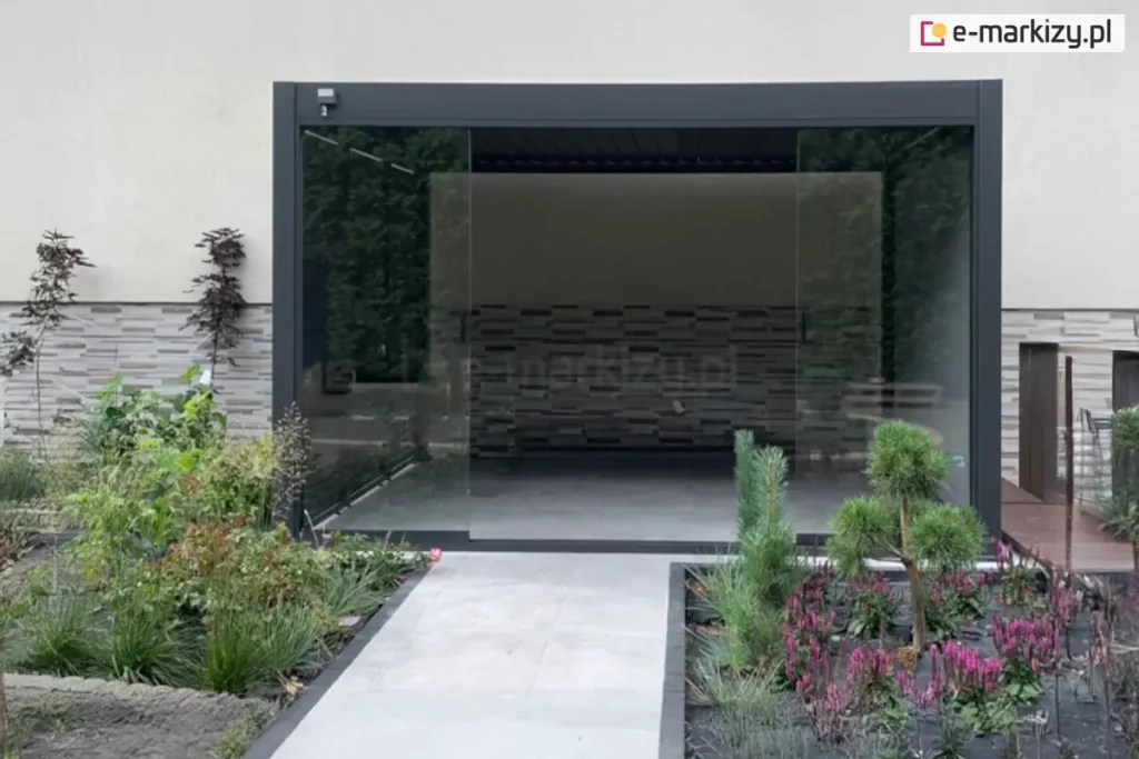 Antracytowa pergola tarasowa Marlo zamontowana jako przedłużenie nowoczesnego budynku, wyposażona w system przesuwnych paneli szklanych tworzących pełną zabudowę boku. Aluminiowa konstrukcja o matowym wykończeniu posiada dach z ruchomych lamel, które na zdjęciu są ustawione w pozycji zamkniętej. Frontowa ściana pergoli składa się z dużych, bezramowych tafli szkła, z których środkowa część jest rozsunięta, tworząc przejście na taras. Ściana po lewej stronie osłonięta została panelem żaluzjowym w kolorze antracytowym, a na prawej ścianie pergoli zamocowano roletę screen. Wewnątrz zabudowanej przestrzeni widoczna jest ozdobna ściana o strukturze szarych, poziomych kamieni lub klocków. Całość osadzona jest na jasnoszarym, gładkim podłożu, które płynnie przechodzi w ogrodową ścieżkę. Przed pergolą zaaranżowano nowoczesny ogród z niskimi nasadzeniami, fioletowymi kwiatami, ozdobnymi trawami oraz niewielkimi iglakami, otoczony ciemnym obrzeżem. Po prawej stronie widoczny jest fragment drewnianego podestu oraz wejście do budynku, a elewacja powyżej pergoli jest gładka i utrzymana w jasnej kolorystyce.