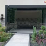 Antracytowa pergola tarasowa Marlo zamontowana jako przedłużenie nowoczesnego budynku, wyposażona w system przesuwnych paneli szklanych tworzących pełną zabudowę boku. Aluminiowa konstrukcja o matowym wykończeniu posiada dach z ruchomych lamel, które na zdjęciu są ustawione w pozycji zamkniętej. Frontowa ściana pergoli składa się z dużych, bezramowych tafli szkła, z których środkowa część jest rozsunięta, tworząc przejście na taras. Ściana po lewej stronie osłonięta została panelem żaluzjowym w kolorze antracytowym, a na prawej ścianie pergoli zamocowano roletę screen. Wewnątrz zabudowanej przestrzeni widoczna jest ozdobna ściana o strukturze szarych, poziomych kamieni lub klocków. Całość osadzona jest na jasnoszarym, gładkim podłożu, które płynnie przechodzi w ogrodową ścieżkę. Przed pergolą zaaranżowano nowoczesny ogród z niskimi nasadzeniami, fioletowymi kwiatami, ozdobnymi trawami oraz niewielkimi iglakami, otoczony ciemnym obrzeżem. Po prawej stronie widoczny jest fragment drewnianego podestu oraz wejście do budynku, a elewacja powyżej pergoli jest gładka i utrzymana w jasnej kolorystyce.