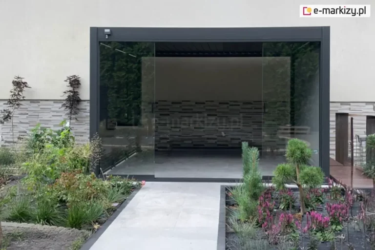 Antracytowa pergola tarasowa Marlo zamontowana jako przedłużenie nowoczesnego budynku, wyposażona w system przesuwnych paneli szklanych tworzących pełną zabudowę boku. Aluminiowa konstrukcja o matowym wykończeniu posiada dach z ruchomych lamel, które na zdjęciu są ustawione w pozycji zamkniętej. Frontowa ściana pergoli składa się z dużych, bezramowych tafli szkła, z których środkowa część jest rozsunięta, tworząc przejście na taras. Ściana po lewej stronie osłonięta została panelem żaluzjowym w kolorze antracytowym, a na prawej ścianie pergoli zamocowano roletę screen. Wewnątrz zabudowanej przestrzeni widoczna jest ozdobna ściana o strukturze szarych, poziomych kamieni lub klocków. Całość osadzona jest na jasnoszarym, gładkim podłożu, które płynnie przechodzi w ogrodową ścieżkę. Przed pergolą zaaranżowano nowoczesny ogród z niskimi nasadzeniami, fioletowymi kwiatami, ozdobnymi trawami oraz niewielkimi iglakami, otoczony ciemnym obrzeżem. Po prawej stronie widoczny jest fragment drewnianego podestu oraz wejście do budynku, a elewacja powyżej pergoli jest gładka i utrzymana w jasnej kolorystyce.