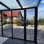 Zdjęcie przedstawia widok z wnętrza nowoczesnej, przeszklonej zabudowy tarasowej na otwartą przestrzeń rekreacyjną i górzysty krajobraz. Konstrukcja oparta jest na systemie szklanych drzwi przesuwnych Sotro w wersji ramowej, gdzie każdy panel ujęty jest w smukłe profile aluminiowe o matowym wykończeniu w kolorze ciemnego antracytu. Wielkoformatowe pakiety szyb zapewniają niezakłócony widok na wyłożony szarą kostką brukową taras z nowoczesnymi, betonowymi siedziskami oraz na dalszy plan z domami o czerwonych dachach i zalesionymi zboczami gór pod błękitnym niebem. Zadaszenie konstrukcji również wykonane jest z przezroczystych paneli szklanych wspartych na systemowych krokwiach, co maksymalnie doświetla wnętrze wykończone dużymi płytami podłogowymi w dwóch odcieniach szarości.