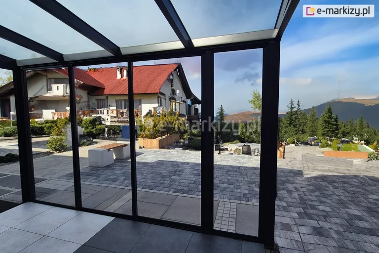 Zdjęcie przedstawia widok z wnętrza nowoczesnej, przeszklonej zabudowy tarasowej na otwartą przestrzeń rekreacyjną i górzysty krajobraz. Konstrukcja oparta jest na systemie szklanych drzwi przesuwnych Sotro w wersji ramowej, gdzie każdy panel ujęty jest w smukłe profile aluminiowe o matowym wykończeniu w kolorze ciemnego antracytu. Wielkoformatowe pakiety szyb zapewniają niezakłócony widok na wyłożony szarą kostką brukową taras z nowoczesnymi, betonowymi siedziskami oraz na dalszy plan z domami o czerwonych dachach i zalesionymi zboczami gór pod błękitnym niebem. Zadaszenie konstrukcji również wykonane jest z przezroczystych paneli szklanych wspartych na systemowych krokwiach, co maksymalnie doświetla wnętrze wykończone dużymi płytami podłogowymi w dwóch odcieniach szarości.