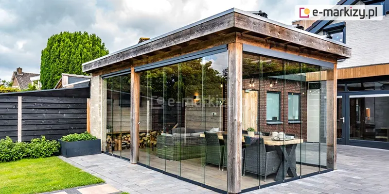 Nowoczesna altana ogrodowa o konstrukcji drewnianej, w której zastosowano system szklanych ścian przesuwnych Auris. Konstrukcja nośna składa się z grubych, surowych belek drewnianych oraz płaskiego dachu z szerokim okapem wykończonym deskami. Przestrzeń między słupami została całkowicie wypełniona bezramowymi panelami z przezroczystego szkła hartowanego, które są osadzone w dyskretnych, ciemnoantracytowych prowadnicach dolnych i górnych. Wewnątrz altany znajduje się strefa wypoczynkowa z szarą sofą technorattanową oraz duży drewniany stół z krzesłami, a na tylnej ścianie widoczne jest składowane drewno kominkowe. Szklane tafle są ustawione w sposób nachodzący na siebie, co tworzy szczelną, a jednocześnie niemal niewidoczną barierę ochronną. Na zewnątrz widoczny jest fragment zadbanego trawnika, ciemnoszary płot lamelowy oraz elewacja budynku z czerwonej cegły, co podkreśla integrację nowoczesnego systemu szklenia z naturalnymi materiałami i architekturą ogrodu.