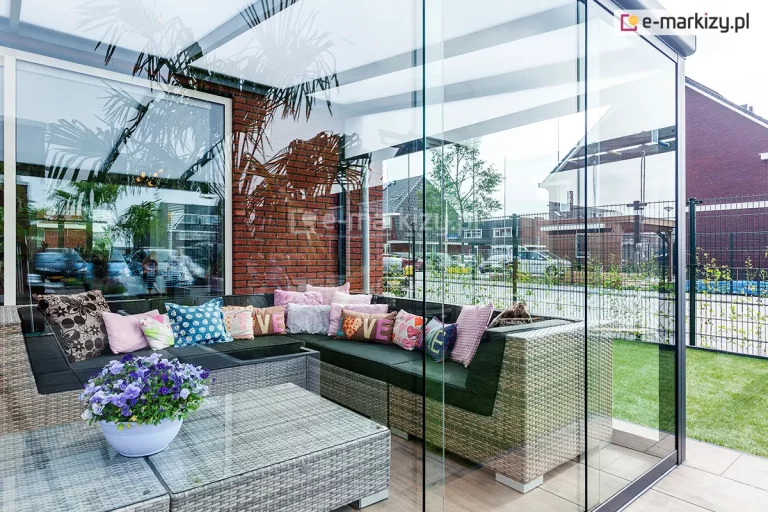 Przestrzeń ogrodu zimowego ukazana z zewnątrz, prezentująca system szklanych ścian przesuwnych Auris w narożnym układzie. Tafle szkła są pozbawione pionowych ram, co daje efekt jednolitej szklanej tafli. Wewnątrz znajduje się wygodna, technorattanowa sofa z licznymi kolorowymi poduszkami o różnych wzorach oraz stolik kawowy z blatem z hartowanego szkła, na którym stoi doniczka z fioletowymi bratkami. Przez szyby widoczna jest elewacja z czerwonej cegły, zaparkowane samochody oraz osiedle domów jednorodzinnych z ogródkami i ogrodzeniami panelowymi.