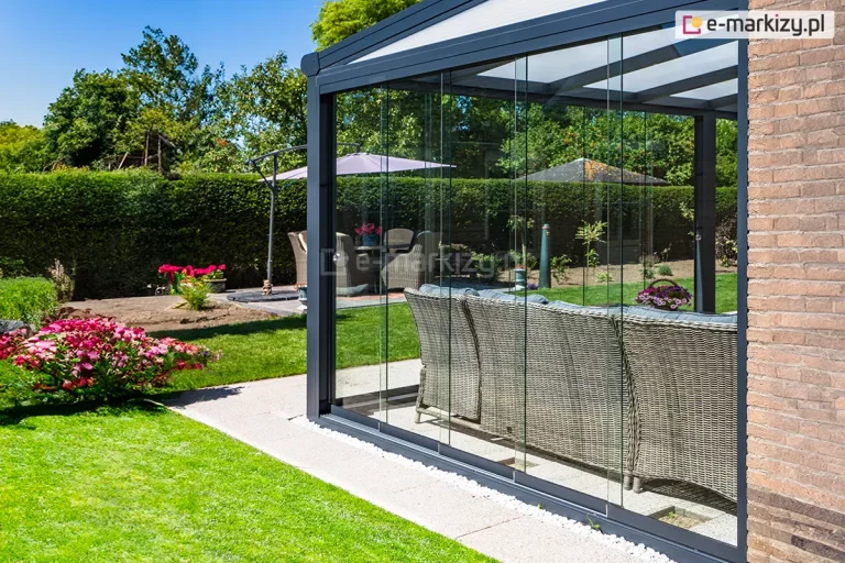 Szklana zabudowa tarasu w systemie bezramowym Auris, zintegrowana z aluminiowym zadaszeniem Lunaro w kolorze antracytowym. Zdjęcie przedstawia narożną sekcję ogrodu letniego, gdzie transparentne panele szklane są zamontowane na wielotorowej prowadnicy dolnej, osadzonej na jasnym pasie żwirowym. Konstrukcja przylega do elewacji budynku z jasnobrązowej cegły. Wewnątrz przeszklonej strefy widoczny jest tył dużej sofy z szarego technorattanu. Na zewnątrz rozpościera się zadbany ogród z zielonym trawnikiem, formowanym żywopłotem oraz kwitnącymi krzewami w kolorze intensywnego różu. W tle widoczny jest drugi zestaw mebli ogrodowych pod parasolem.
