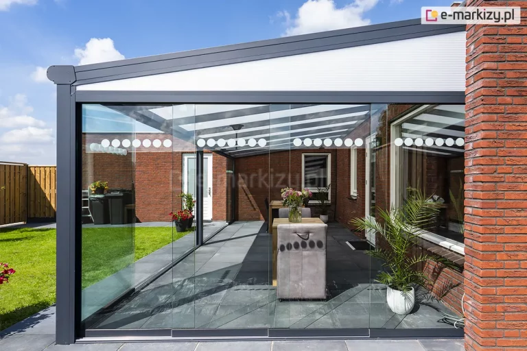 Boczne ujęcie nowoczesnego zadaszenia tarasowego Lunaro w kolorze antracytowym, które zostało wyposażone w system bezramowych szklanych ścian przesuwnych Auris. Konstrukcja posiada jednospadowy dach z wypełnieniem z białego, półprzezroczystego poliwęglanu, zamontowany do budynku z czerwonej cegły. Powierzchnia pomiędzy spadkiem dachu a bocznymi przeszkleniami została wypełniona klinem z poliwęglanem komorowym w kolorze mlecznym. Szklane panele, chroniące wnętrze tarasu, ozdobione są charakterystycznym poziomym pasem białych, okrągłych naklejek, które pełnią funkcję ostrzegawczą i dekoracyjną. Wewnątrz przeszklonej przestrzeni, na tarasie wyłożonym ciemnoszarymi płytami, widoczna jest aranżacja z drewnianymi stołami oraz fioletowymi, tapicerowanymi fotelami, a także doniczkowe rośliny ozdobne. W tle, za przeszkleniem, rozpościera się widok na zielony trawnik, drewniane ogrodzenie oraz błękitne niebo z lekkim zachmurzeniem.