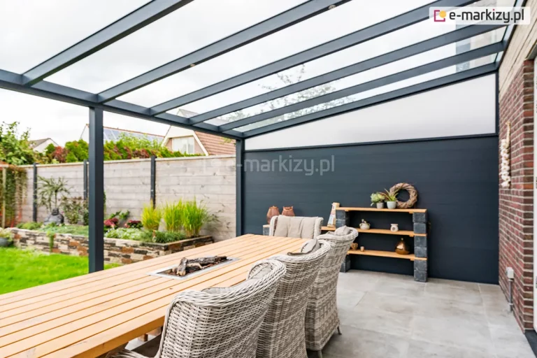 Nowoczesne szklane zadaszenie tarasowe model Terano o konstrukcji wykonanej z profili aluminiowych w kolorze antracytowym. Zadaszenie posiada jednospadowy dach pokryty transparentnymi panelami szklanymi, które są osadzone na równolegle rozmieszczonych krokwiach. Konstrukcja wsparta jest na pionowym słupie nośnym o kwadratowym przekroju. Jedna z bocznych ścian tarasu została zabudowana pełną osłoną w formie ciemnoszarej palisady zintegrowanej z górnym trójkątnym wypełnieniem z poliwęglanu o mlecznym zabarwieniu. Pod zadaszeniem znajduje się strefa wypoczynkowa z dużym drewnianym stołem wyposażonym w centralne palenisko oraz plecionymi fotelami technorattanowymi w kolorze szarym. Podłoże tarasu wyłożone jest dużymi, jasnymi płytami gresowymi. W tle widoczny jest ogród z kamiennymi murkami oporowymi, roślinnością ozdobną oraz drewnianym ogrodzeniem posesji.