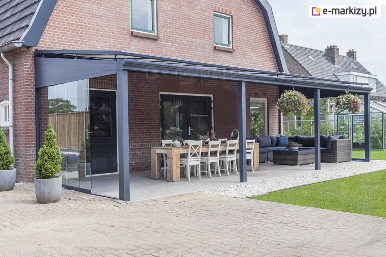 Rozbudowane szklane zadaszenie tarasu model Terano, oparte na czterech solidnych profilach aluminiowych w kolorze antracytowym. Konstrukcja została zamontowana wzdłuż elewacji piętrowego budynku wykonanego z czerwonej cegły, przykrytego ciemnym dachem o charakterystycznym załamaniu. Zadaszenie posiada wypełnienie z przezroczystych paneli szklanych, które wpuszczają naturalne światło na całą powierzchnię tarasu. Lewa strona konstrukcji została zabudowana systemem przesuwnych drzwi szklanych, tworząc zamkniętą, transparentną ścianę boczną. Pod zadaszeniem wydzielono dwie strefy funkcjonalne: jadalnianą z długim, drewnianym stołem i białymi krzesłami oraz wypoczynkową z dużym narożnikiem ogrodowym z ciemnoszarymi poduszkami i niskim stolikiem. Z belek konstrukcyjnych zwisają dekoracyjne donice z kwitnącymi, różowo-fioletowymi kwiatami. Podłoga tarasu jest wybetonowana, wykończona gładką powierzchnią w jasnym kolorze, a jej krawędź oddzielona jest od trawnika pasem jasnego żwiru. W tle widoczny jest zadbany ogród oraz sąsiednia zabudowa z szklarnią.