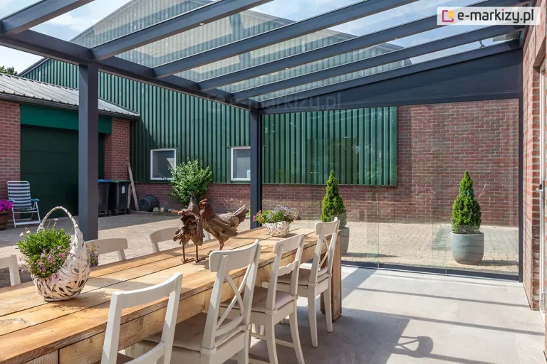 Nowoczesne zadaszenie tarasu o konstrukcji aluminiowej w kolorze ciemnoszarym, wyposażone w przezroczyste panele szklane na dachu. Konstrukcja opiera się na pionowych słupach o kwadratowym przekroju, które podtrzymują poziome belki oraz skośne krokwie dachowe. System zadaszenia jest zamontowany bezpośrednio do ściany budynku wykonanej z czerwonej cegły. Na bocznej ścianie konstrukcji zainstalowano przesuwne panele szklane, które tworzą transparentną barierę chroniącą przed wiatrem, zachowując przy tym pełną widoczność na otoczenie. Pod zadaszeniem znajduje się strefa jadalniana z dużym, masywnym stołem wykonanym z jasnego drewna, przy którym ustawiono białe, klasyczne krzesła z oparciami. Na stole umieszczono dekoracyjne wiklinowe kosze z fioletowymi kwiatami oraz ozdobne figurki ptaków. Podłoga tarasu wyłożona jest jasnoszarymi płytami wielkoformatowymi. W tle widoczny jest wybrukowany podjazd, zielony budynek gospodarczy z metalowej blachy falistej oraz rośliny doniczkowe w postaci formowanych krzewów.