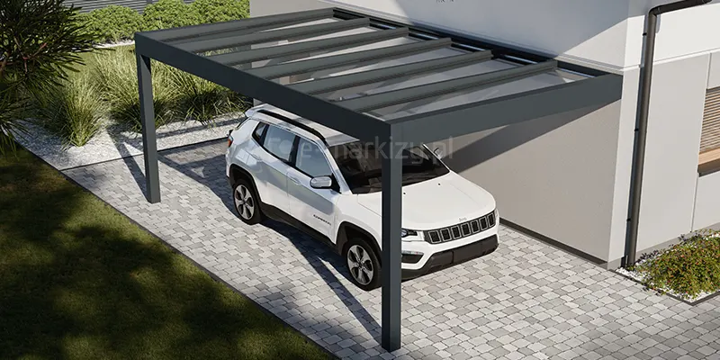 Nowoczesna konstrukcja szklanego carportu Spekta Glass, zaadaptowanego jako zadaszenie podjazdowe przy ścianie budynku. Konstrukcja nośna została wykonana z grubych profili aluminiowych w kolorze antracytowym, tworzących ramę dachu wspartą na dwóch solidnych słupach od strony zewnętrznej. Dach jest w całości przeszklony i posiada lekki spadek, co umożliwia skuteczne odprowadzanie wody opadowej, jednocześnie zapewniając pełną widoczność. Pod zadaszeniem zaparkowany jest biały samochód osobowy typu SUV, co obrazuje funkcjonalne zastosowanie systemu jako wiaty garażowej. Podłoże pod konstrukcją wyłożone jest betonową kostką brukową w odcieniach szarości, ułożoną w regularny wzór. W tle widoczna jest nowoczesna elewacja budynku z ciemną rynną oraz niska roślinność ogrodowa z ozdobnymi trawami i krzewami na tle białego gresu, co podkreśla techniczny i estetyczny charakter instalacji.