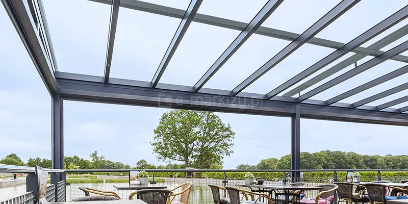 Szklany dach nad tarasem ze stolikami z widokiem na rzekę oraz drzewa w oddali.