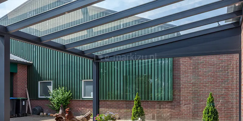 Zdjęcie prezentuje widok od wewnątrz na konstrukcję szklanego dachu zadaszenia Terano. Aluminiowe krokwie w kolorze antracytowym podtrzymują tafle przezroczystego szkła, a w każdym profilu zamontowano punktowe diody LED. Konstrukcja jest zamontowana do budynku, którego elewację stanowi połączenie czerwonej cegły oraz zielonej blachy trapezowej. Widoczne są również słupy nośne podtrzymujące przednią część dachu. W tle znajduje się podwórze z ozdobnymi figurkami i niską roślinnością.