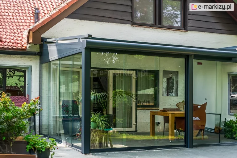 Szklany ogród letni zbudowany na bazie systemu Terano, przylegający do białej elewacji budynku. Aluminiowa konstrukcja w kolorze antracytowym posiada szklany dach oraz przesuwne ściany boczne wykonane z bezramowego szkła. Wewnątrz widoczne są rośliny doniczkowe, drewniany stół oraz nowoczesne fotele. Budynek charakteryzuje się tradycyjną dachówką ceramiczną i ciemnymi obramowaniami okien. Przed konstrukcją znajduje się betonowy podest tarasowy z donicami obsadzonymi różnorodnymi roślinami.